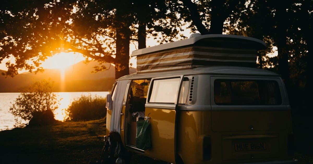 A caravan parked on a gravel driveway beside a weatherboard rural Australian house, with a kelpie sitting on the verandah in late afternoon light.