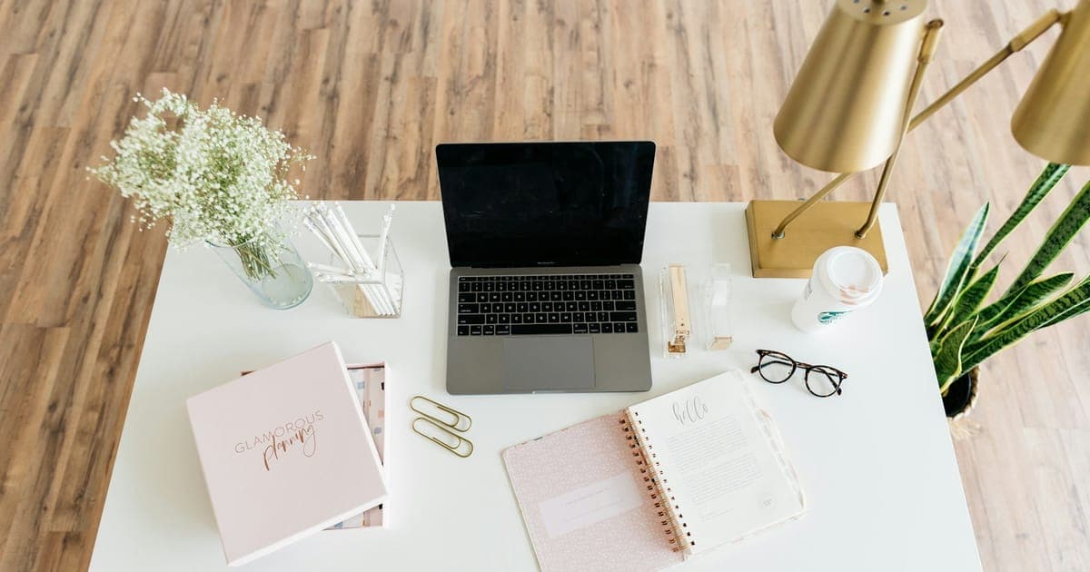An overhead view of a white desk with an open laptop, a spiral notebook, reading glasses, a coffee cup and a potted plant