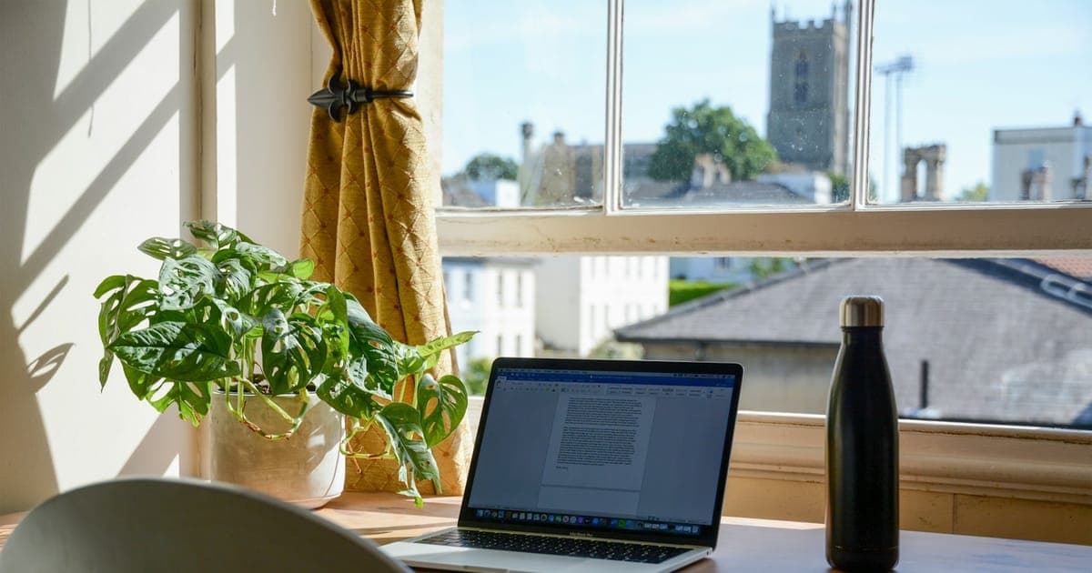 A woman on a phone call at a kitchen bench with a notebook open in front of her, giving a reference for a house sitter while a cat sits on the stool beside her.