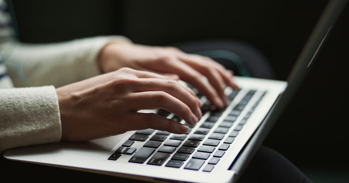 A homeowner at a kitchen bench writing a house sitting listing on a laptop, with a kelpie cross lying on the floor beside the stool.